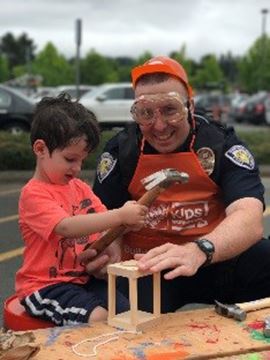 BPD officer, wearing safety goggles, helps young child hammering on a wooden craft project.